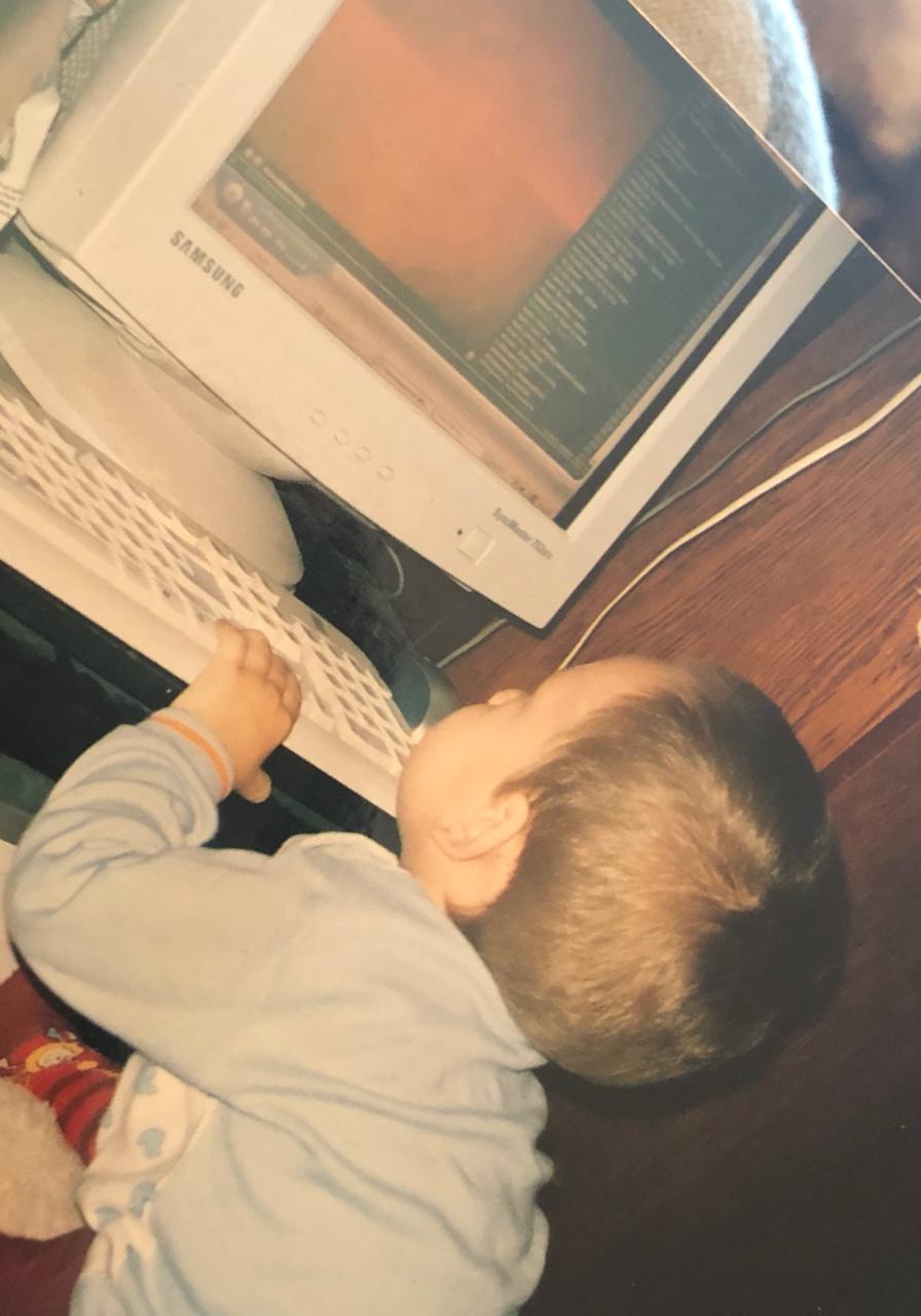 Toddler in blue pajamas sitting at a desk, hands on a keyboard in front of a Samsung SyncMaster CRT monitor