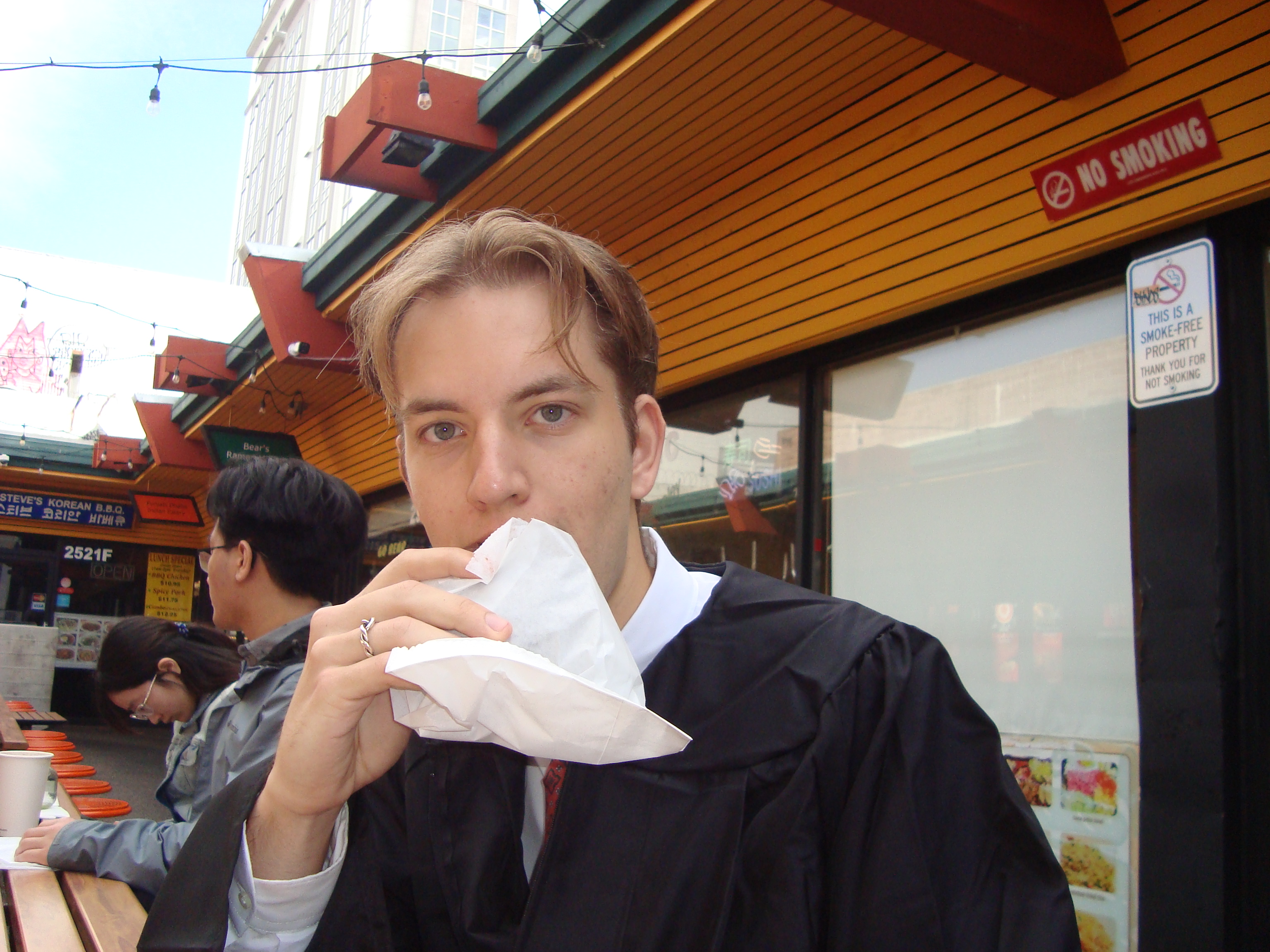 Graduation-day photograph outdoors, holding a paper-wrapped donut in front of food stalls